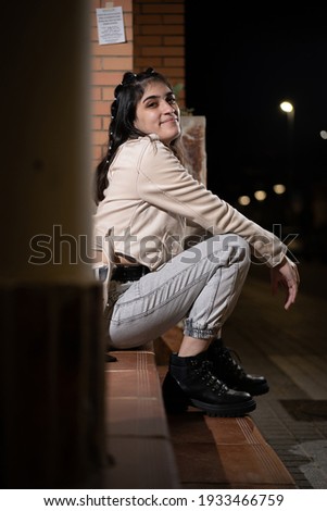 Caucasian white young woman with black hair and stylish urban clothes, white conc, black and white striped t-shirt, gray jeans and boots, sitting posing on some stairs at night with flash light