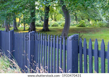 A black wooden picket fence with green grass, large trees, and lush shrubs in a garden. The summer scene is vibrant and colorful. The sun is shining on the dark palings giving a warm glow to the wood. Royalty-Free Stock Photo #1933470431