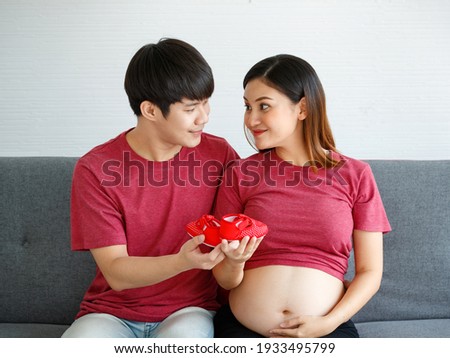 Portrait of a lovely young family couple. A man and a pregnant woman wearing a red T-shirt sitting on a couch smiling and looking at each other holding a red baby shoe.