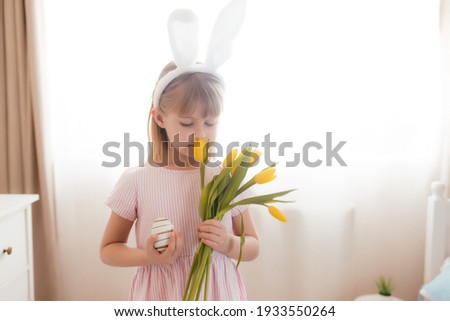 Easter concept. Small smiling girl in white fluffy bunny ears and pink dress holding yellow tulip flowers and colorful egg. Room interior. High quality photo