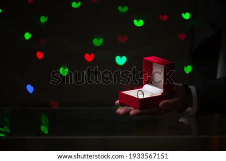 Valentine's Day engagement. A man hand holds a wedding ring in a red box. Behind on a dark background red and green hearts
