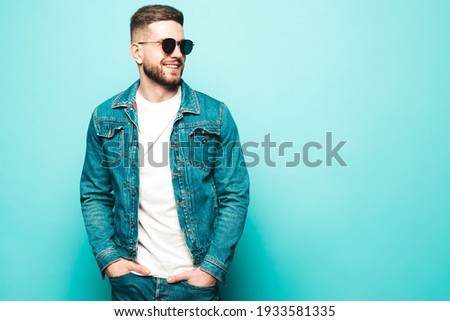 Portrait of handsome smiling stylish hipster lambersexual model.Man dressed in jacket and jeans. Fashion male posing near blue wall in studio in sunglasses Royalty-Free Stock Photo #1933581335