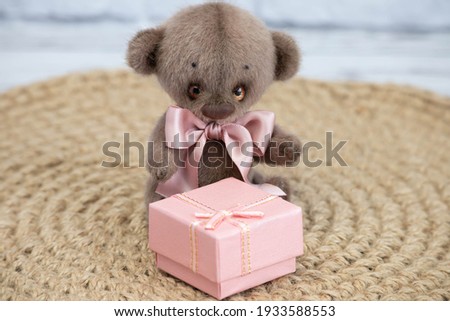 A cute teddy bear with a pink bow sits on a wicker background. There is a beautiful gift box next to it.