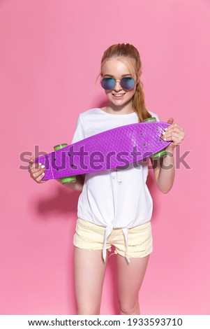 Cheerful young blonde girl with a skateboard on the pink background.