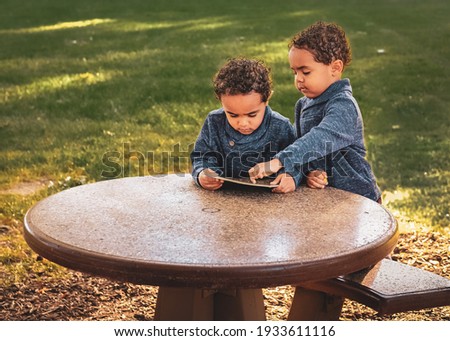 African-American twin toddler boys sitting at table in park in fall with a book at sunset; one boy is pointing to page