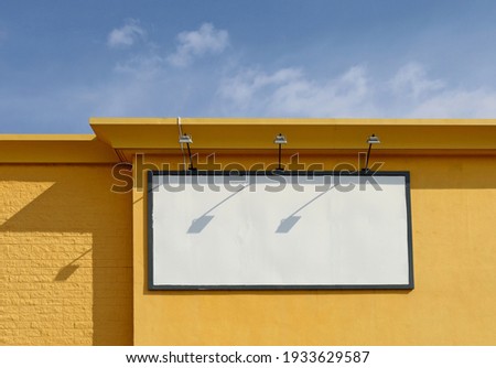 Blank advertising board, with spotlights above, on top of a yellow building  wall. Blue sky above. Copy space