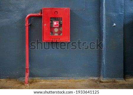 Fire hose in red box. Pipe roll for fire emergency in red metal cabinet on gray painted concrete wall with tiled floor as part of firefighting system of industrial production plant with copyspace.