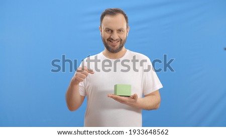 Young man with a beard in front of a blue background is showing the green little box in his left hand with his right index finger and looking at the screen and laughing. 