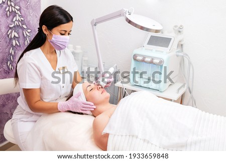 New normal. A cosmetologist in rubber gloves and medical mask does a rf-lifting procedure on the face. In the background, the Rf cosmetology device. Concept of professional cosmetology and health. Royalty-Free Stock Photo #1933659848
