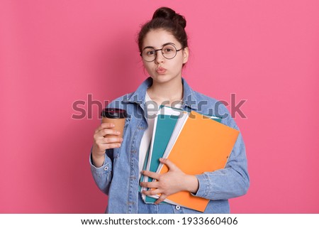 Lovely dark dark haired schoolgirl keeps lips rounded, flirts with somebody at school, wears round spectacles, holds colorful paper folder, disposable cup of coffee or cappuccino.