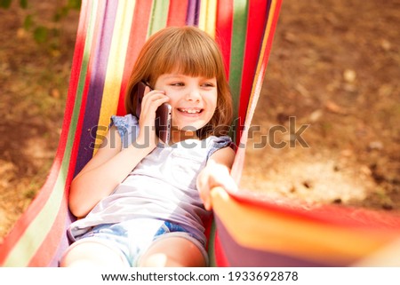 Pretty smiling child girl laying on hammock and talking on mobile phone.