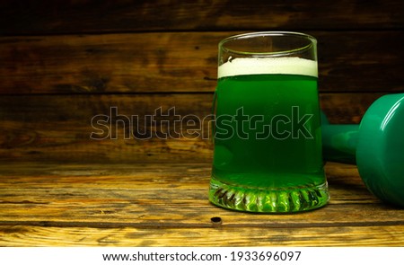 Glass of Irish green beer and heavy dumbbell on wooden background. Healthy fitness gym composition concept for St. Patrick's Day, with copyspace. Cheat day temptation vs sticking to the diet.