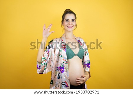 Young beautiful woman pregnant expecting baby wearing pajama over isolated yellow background doing hand symbol