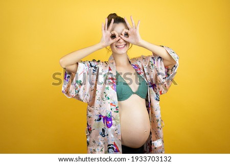 Young beautiful woman pregnant expecting baby wearing pajama over isolated yellow background doing ok gesture shocked with smiling face, eye looking through fingers