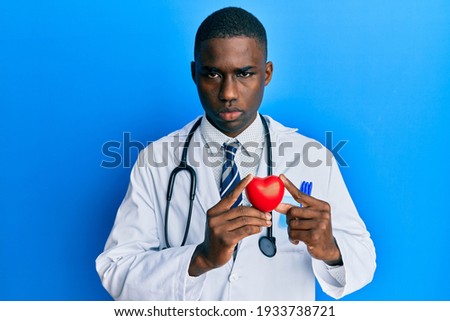 Young african american man wearing doctor uniform holding heart skeptic and nervous, frowning upset because of problem. negative person. 