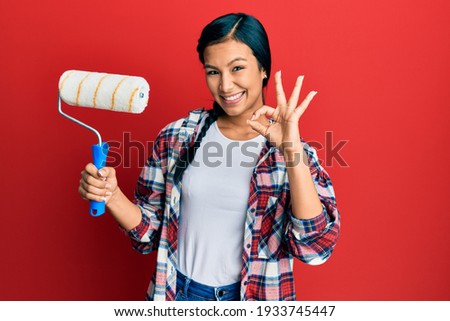 Beautiful hispanic woman holding roller painter doing ok sign with fingers, smiling friendly gesturing excellent symbol 