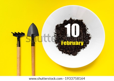 February 10th. Day 10 of month, Calendar date. White plate of soil with a small spatula and rake on yellow background. Winter month, day of the year concept