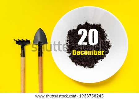 December 20th. Day 20 of month, Calendar date. White plate of soil with a small spatula and rake on yellow background. Winter month, day of the year concept