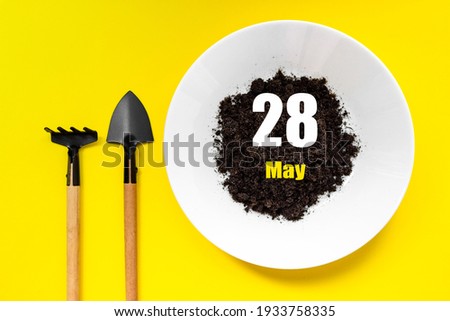 May 28th. Day 28 of month, Calendar date. White plate of soil with a small spatula and rake on yellow background. Spring month, day of the year concept