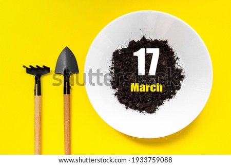 March 17th. Day 17 of month, Calendar date. White plate of soil with a small spatula and rake on yellow background. Spring month, day of the year concept