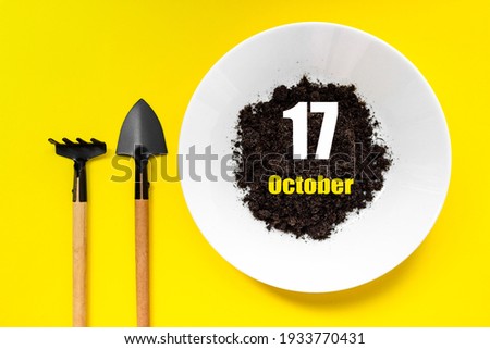 October 17th. Day 17 of month, Calendar date. White plate of soil with a small spatula and rake on yellow background. Autumn month, day of the year concept
