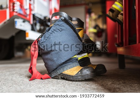Firefighter's shoes and pants and a wardrobe cabinet, fire truck and fireman in the background.