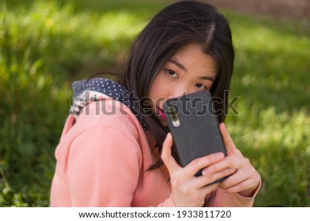 happy Asian woman using mobile phone at city park - young sweet and beautiful Korean girl sitting on green grass networking enjoying social media with hand phone