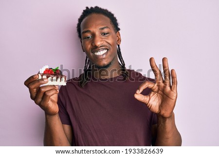 African american man with braids holding chocolate cake doing ok sign with fingers, smiling friendly gesturing excellent symbol 
