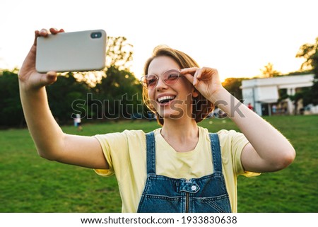 Image of cheerful brunette woman taking selfie photo on cellphone and smiling while resting in summer park