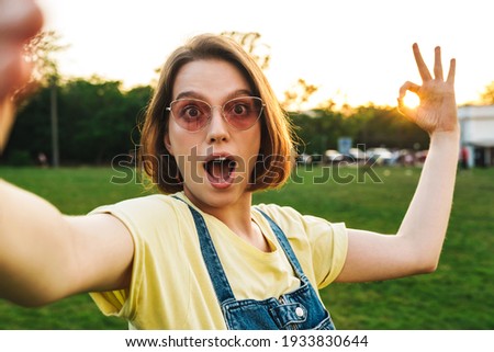 Image of surprised brunette woman taking selfie and showing ok sign while resting in summer park