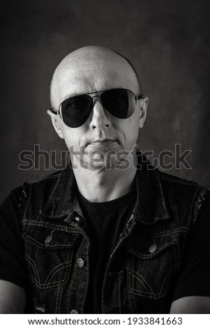 Unshaven, bald middle-aged man in a black T-shirt, denim vest and dark glasses. Black and white portrait. Photo taken in studio
