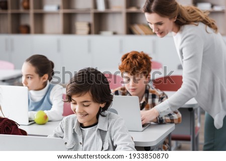 teacher helping schoolboy working on laptop during lesson, blurred background