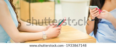 Hands of two young white women holding smartphones sitting at the table outdoors