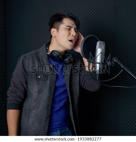 Young asian man with headphones  singing in front of black soundproofing walls. Musicians producing music in professional recording studio.