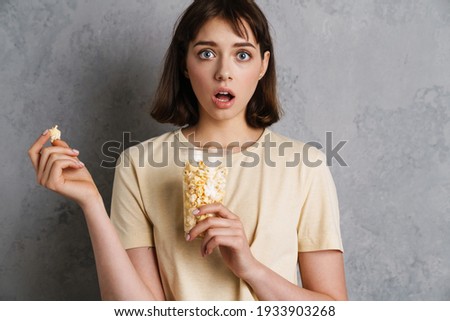 Excited shocked young girl eating popcorn while watching TV isolated over gray background