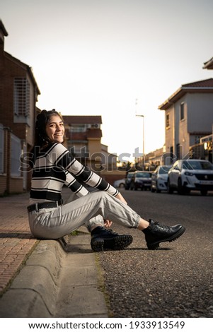 Caucasian young woman in black and white striped t-shirt, black hair, gray jeans and boots, sitting on a curb by the road. Fashion. Style