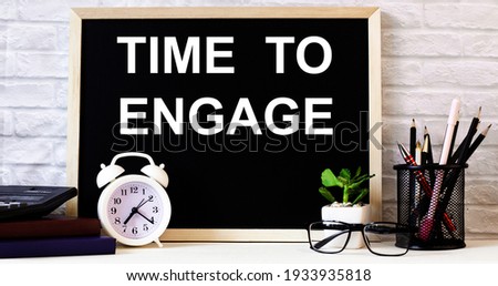 The words TIME TO ENGAGE is written on the chalkboard next to the white alarm clock, glasses, potted plant, and pencils in a stand.
