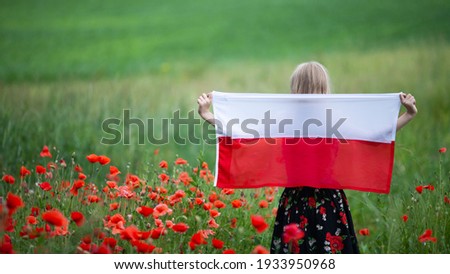 Blond girl holding flag of Poland in the poppy field. Back view. Polish Flag Day. Independence Day. Travel and learn polish language concept. Selective focus. Royalty-Free Stock Photo #1933950968