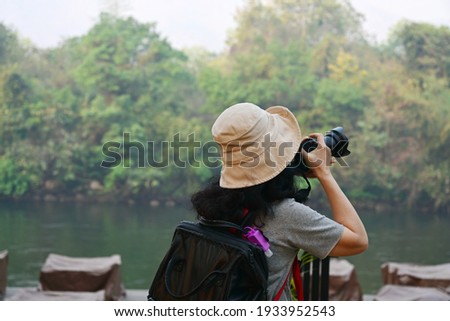 Asian female tourists wearing a cream-colored hat, carrying black backpack, hanging bottles of purple hand sanitizer, taking photos of the nature of the beautiful forest and river on a camera vacation