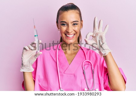 Young hispanic woman wearing doctor stethoscope holding syringe doing ok sign with fingers, smiling friendly gesturing excellent symbol 