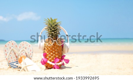 Summer party. Pineapple wearing sunglasses and listen to music with sunblock and sandal on beach and blue sky background. Tropical fashion. Summer Fashion on holiday concept.  Royalty-Free Stock Photo #1933992695