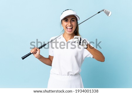 Young hispanic golfer woman over isolated blue wall surprised and pointing front