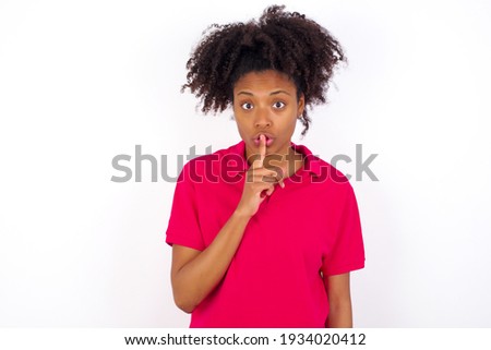 young beautiful African American woman wearing pink t-shirt against white wall makes hush gesture, asks be quiet. Don't tell my secret or not speak too loud, please!