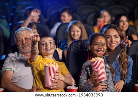 Front view of multiracial kids holding popcorn and sitting on knees of parents. Little african girl with caucasian woman and caucasian boy with senior grandfather. Family, entertainment concept.