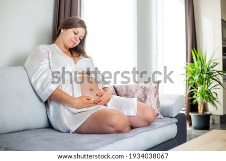 Young pregnant woman sitting on the sofa and enjoying lovely moments of her first pregnancy time