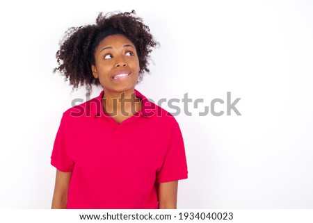 young beautiful African American woman wearing pink t-shirt against white wall with thoughtful expression, looks away keeps hands down bitting his lip thinks about something pleasant.