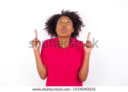 young beautiful African American woman wearing pink t-shirt against white wall being amazed and surprised looking and pointing up with fingers showing something strange.