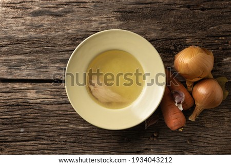 Bowl with clear rich broth. White crockery on an aged wooden table. Vegetables - carrots, onions, garlic, spices