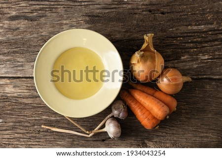 Bowl with clear rich broth. White crockery on an aged wooden table. Vegetables - carrots, onions, garlic, spices