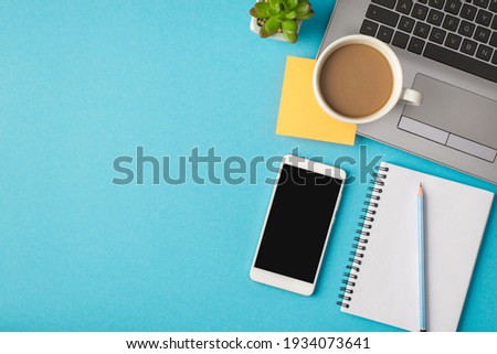 Top view photo of workplace with laptop pencil on notebook yellow sticker smartphone plant and cup of coffee on isolated blue background with blank space
